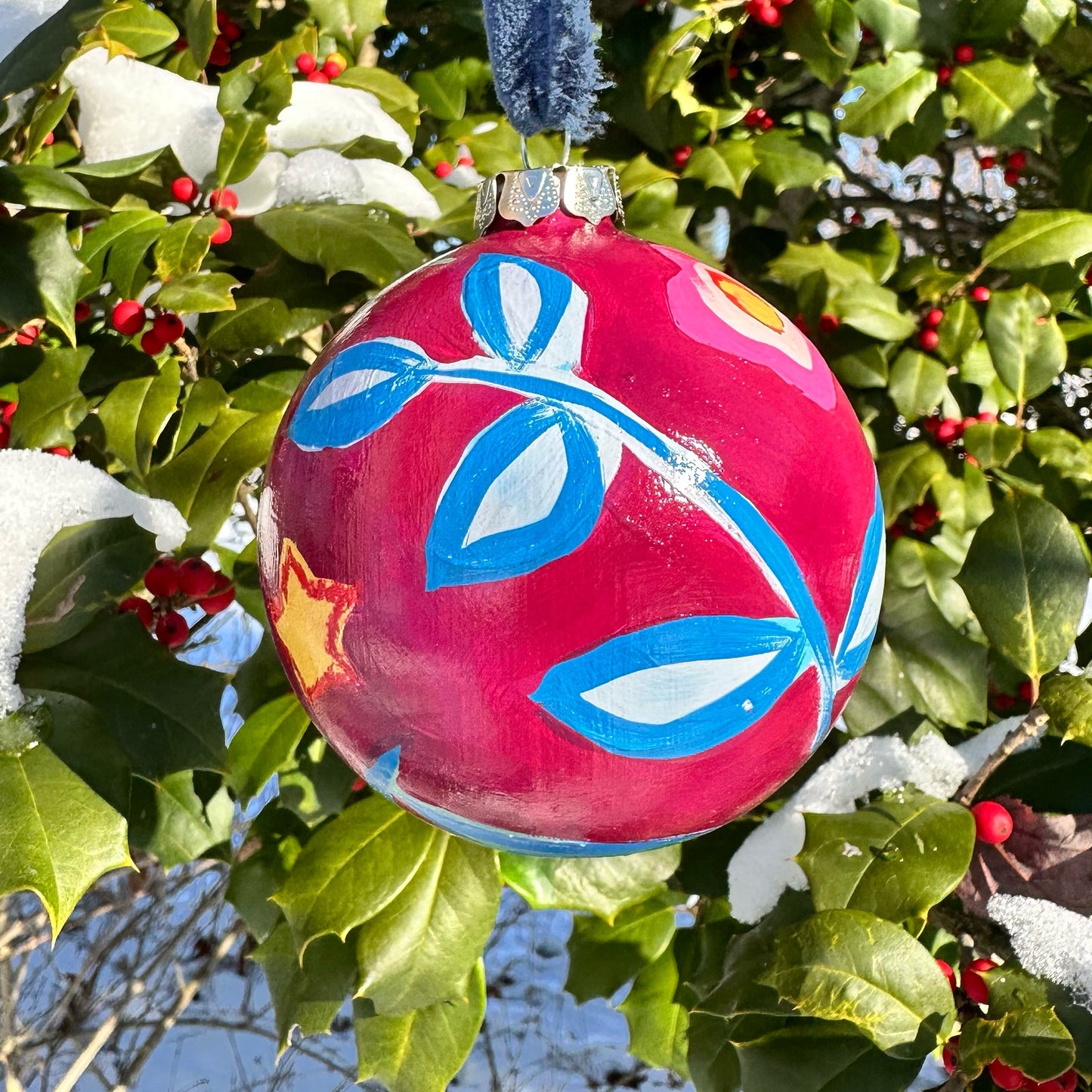 Decorative pink ornament with blue and white designs hanging on a holly bush.