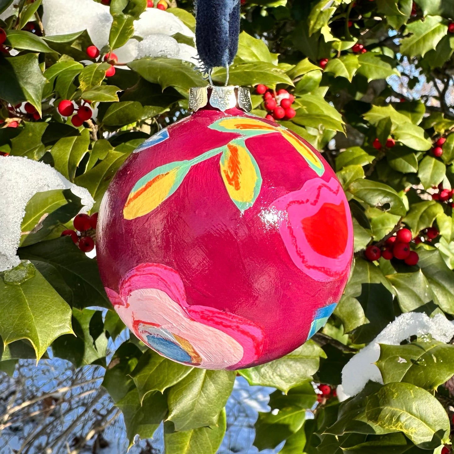 Colorful hand-painted Christmas ornament hanging on a holly branch with berries.